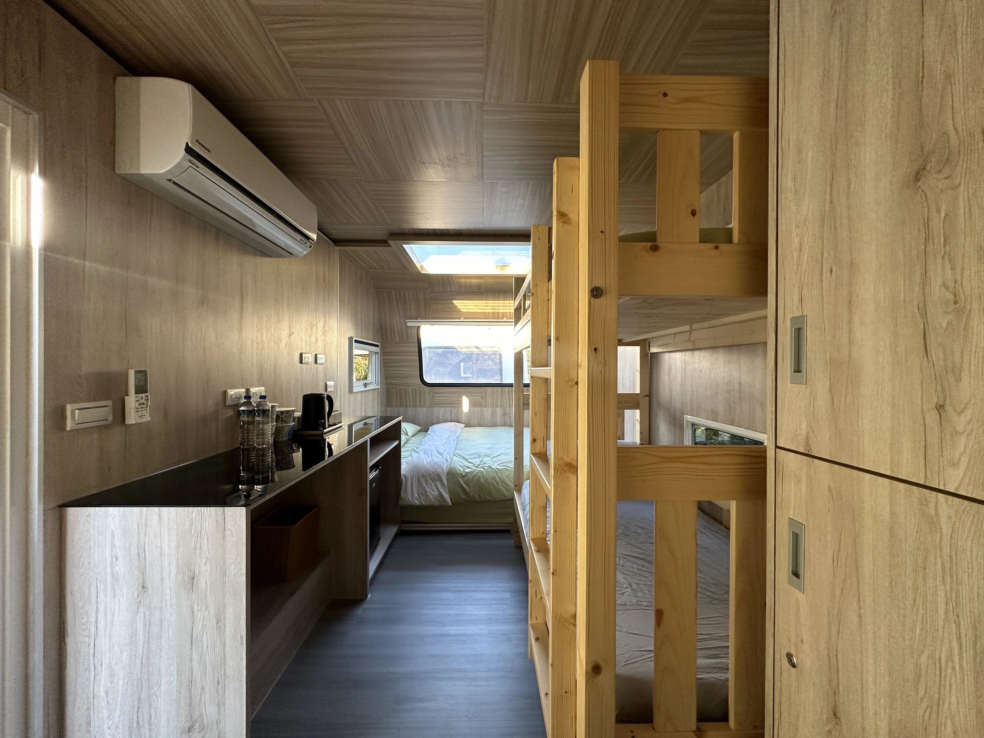 兩張床鋪的房車內部,左邊是一張雙人床,右邊是一個木製上下鋪。房間有木質紋理牆壁和天花板,上方有一個天窗。