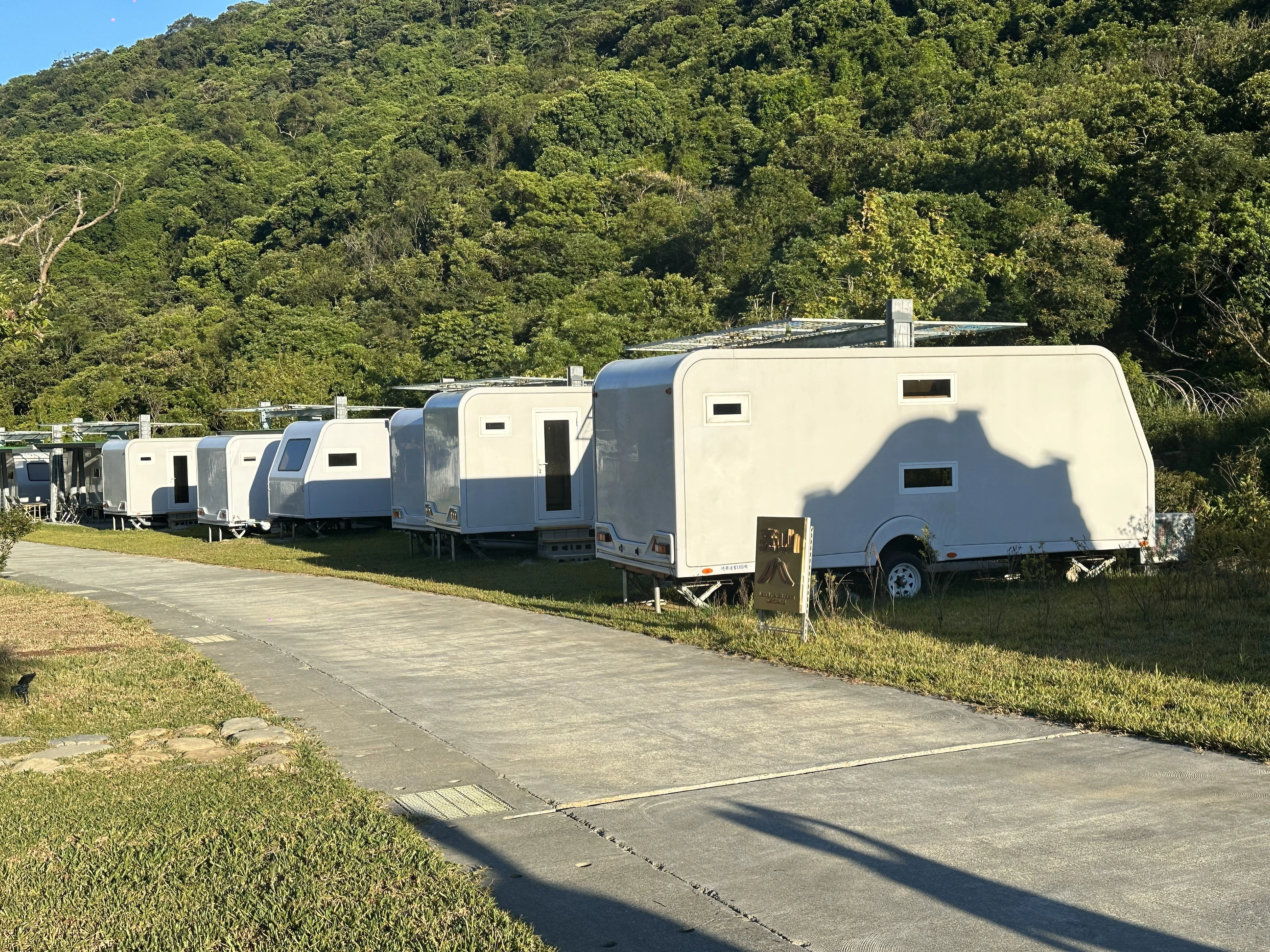 一排白色露營車停在綠色山丘旁的露營區道路上,營造出戶外露營的氣氛