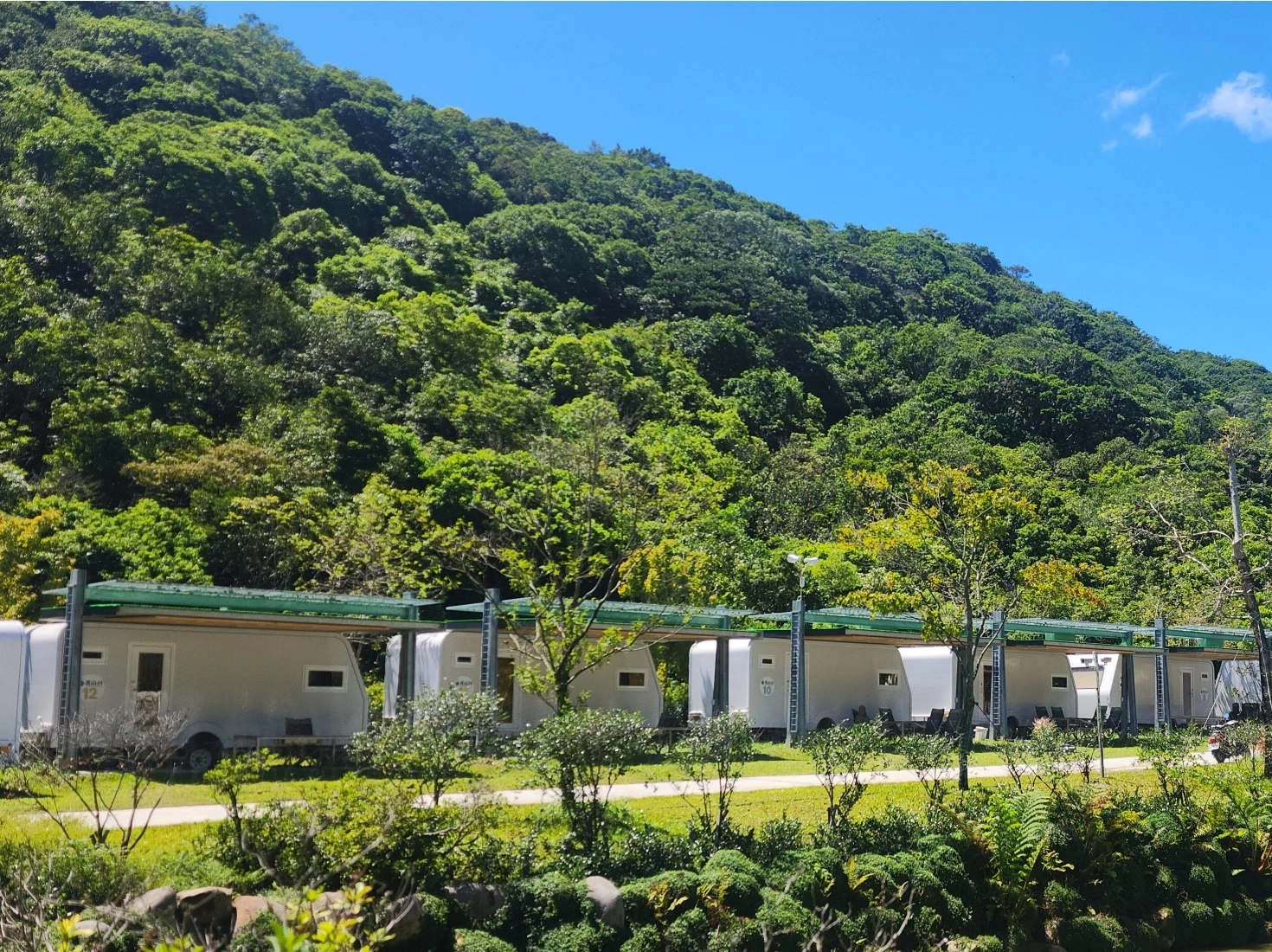 亮山川｜懶人露營｜陽光明媚的藍天下，一排現代化的白色露營車整齊地排列在濃密翠綠的山腳下，周圍環繞著豐富的自然植被