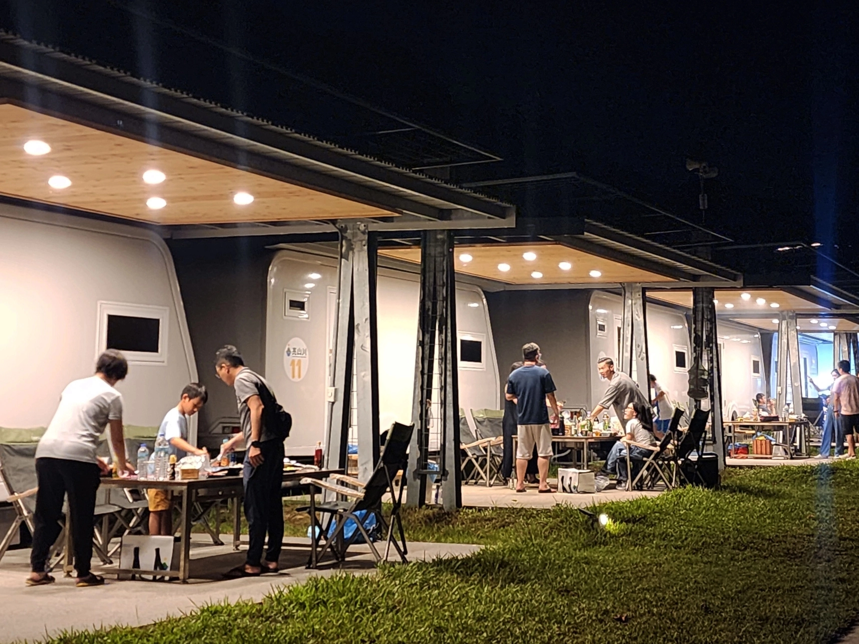 亮山川｜懶人露營｜夜晚草地上排列整齊的露營車，每輛車前設有明亮的木質遮棚與餐桌，多個家庭正悠閒地聚餐與聊天，營造出溫馨熱鬧的戶外度假氛圍